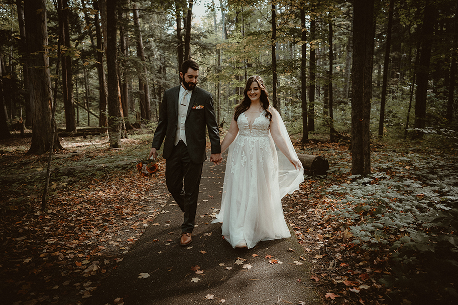 Bride and groom holding hands walking down a path in the woods with autumn leaves on the ground.