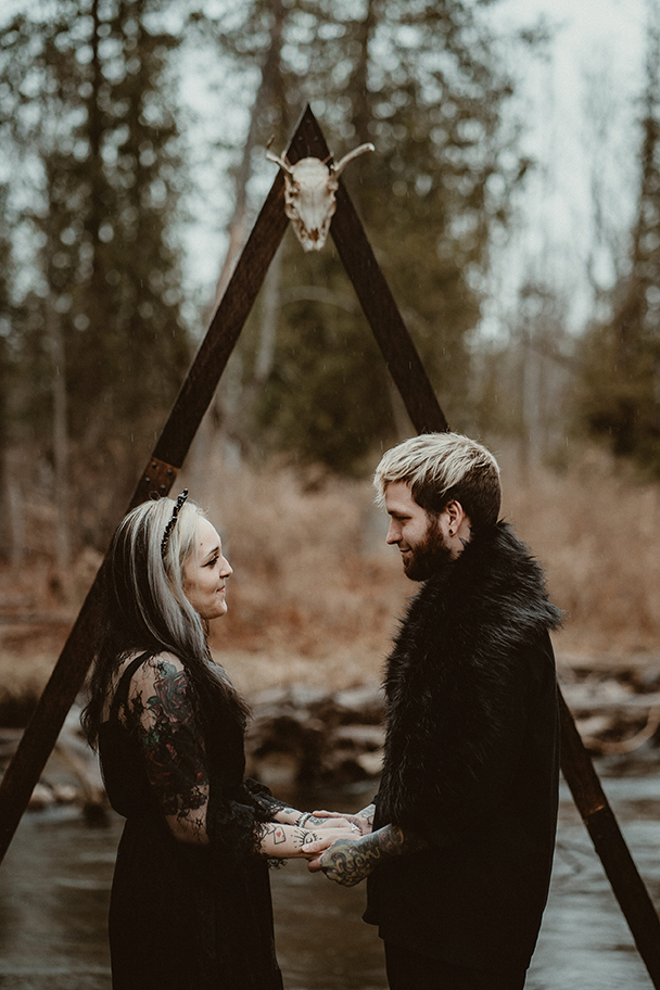 Bride and Groom in black wedding clothes standing together at the ceremony arch with a deer skull at the top.