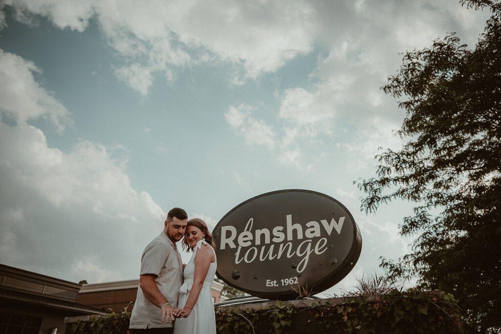 Couple standing in front of the Renshaw Lounge sign outside with cloudy blue skies above.