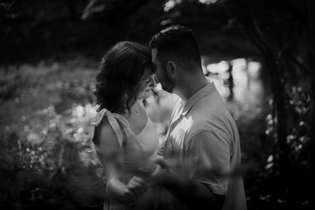 Black and white image of couple holding each other near the river with the sun lighting their faces.