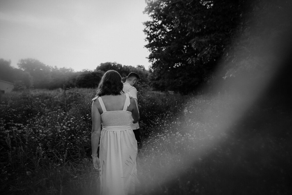 Black and white image of couple walking through a path of wildflowers with him leading her away from the camera.