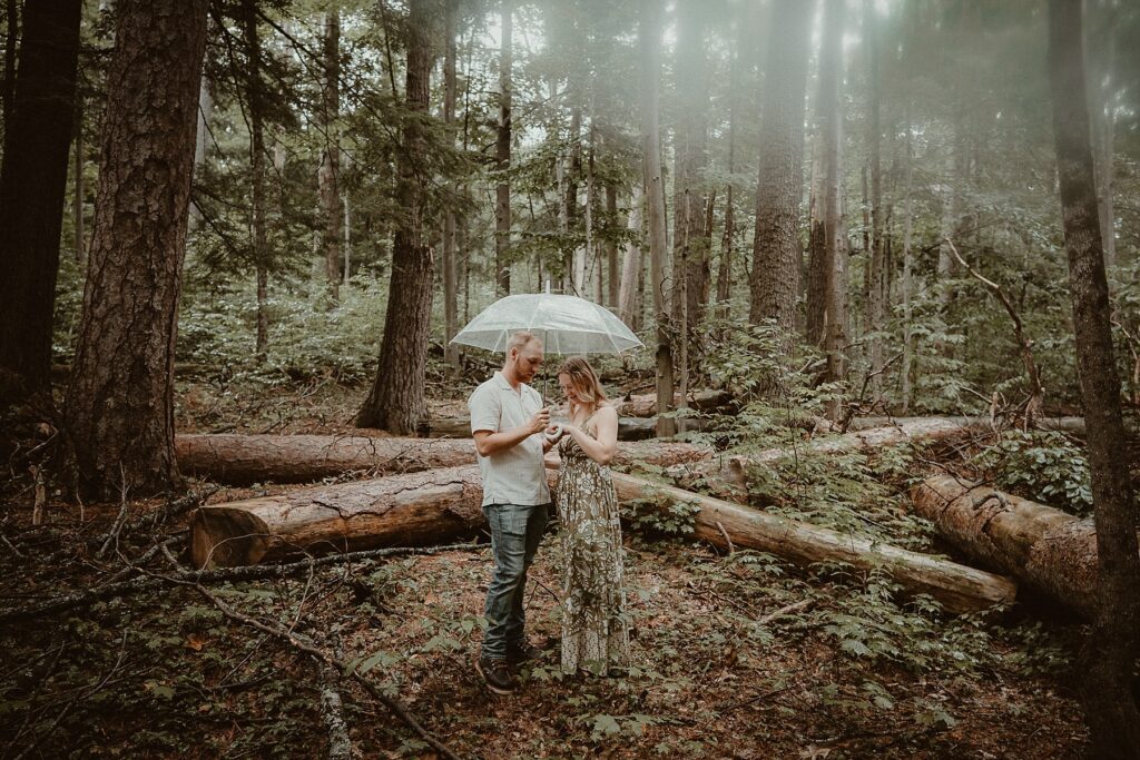 Couple standing together under a clear umbrella in the woods.