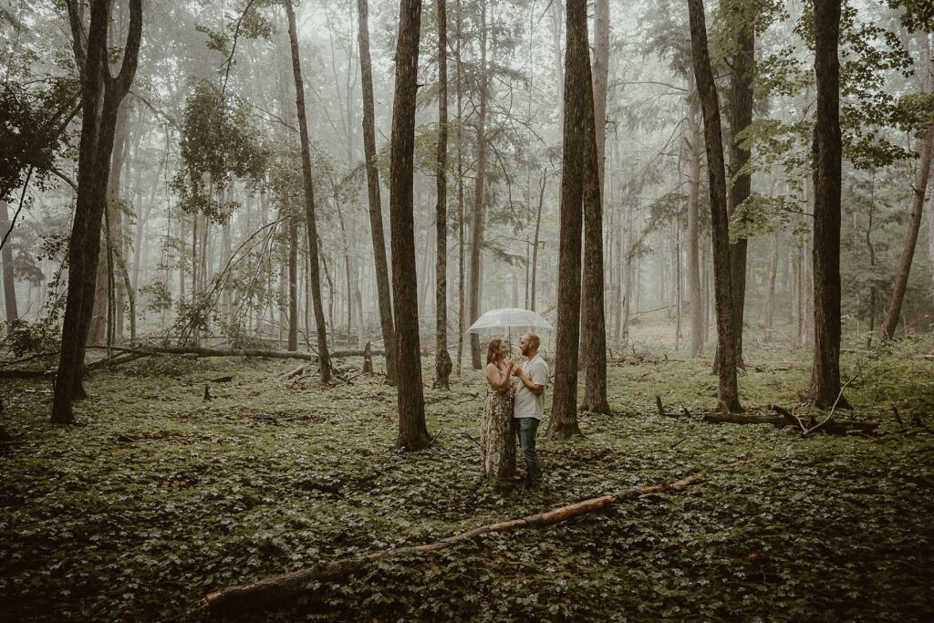 Couple standing together in the rain under a clear umbrella in the woods.