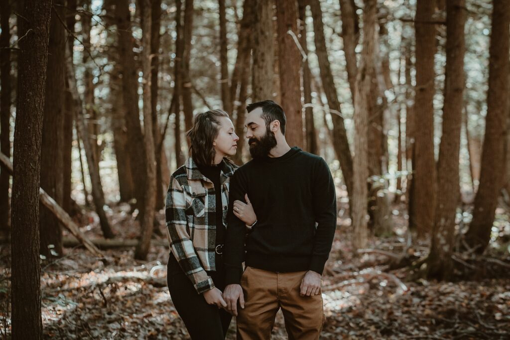 ngaged couple standing beneath towering old-growth pines surrounded by golden autumn leaves.