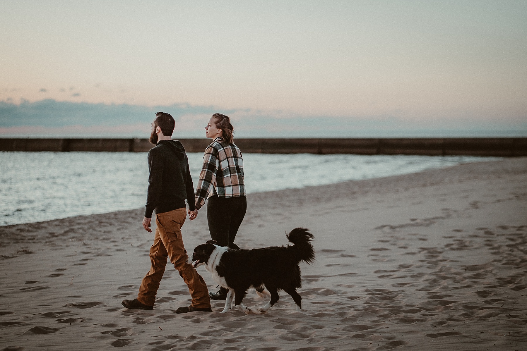Engaged couple walking on the beach of Lake Michigan at sunset with Border Collie between them.