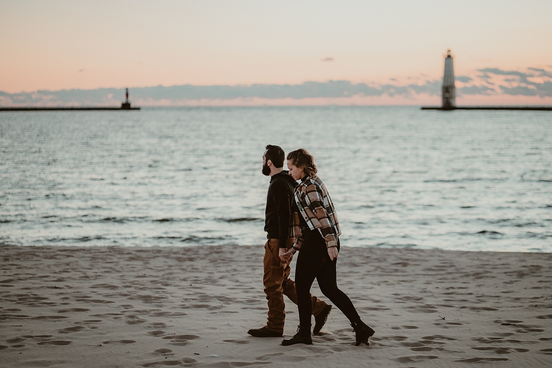 Engaged couple standing on the beach at sunset with Frankfort Light visible on the breakwater behind them.