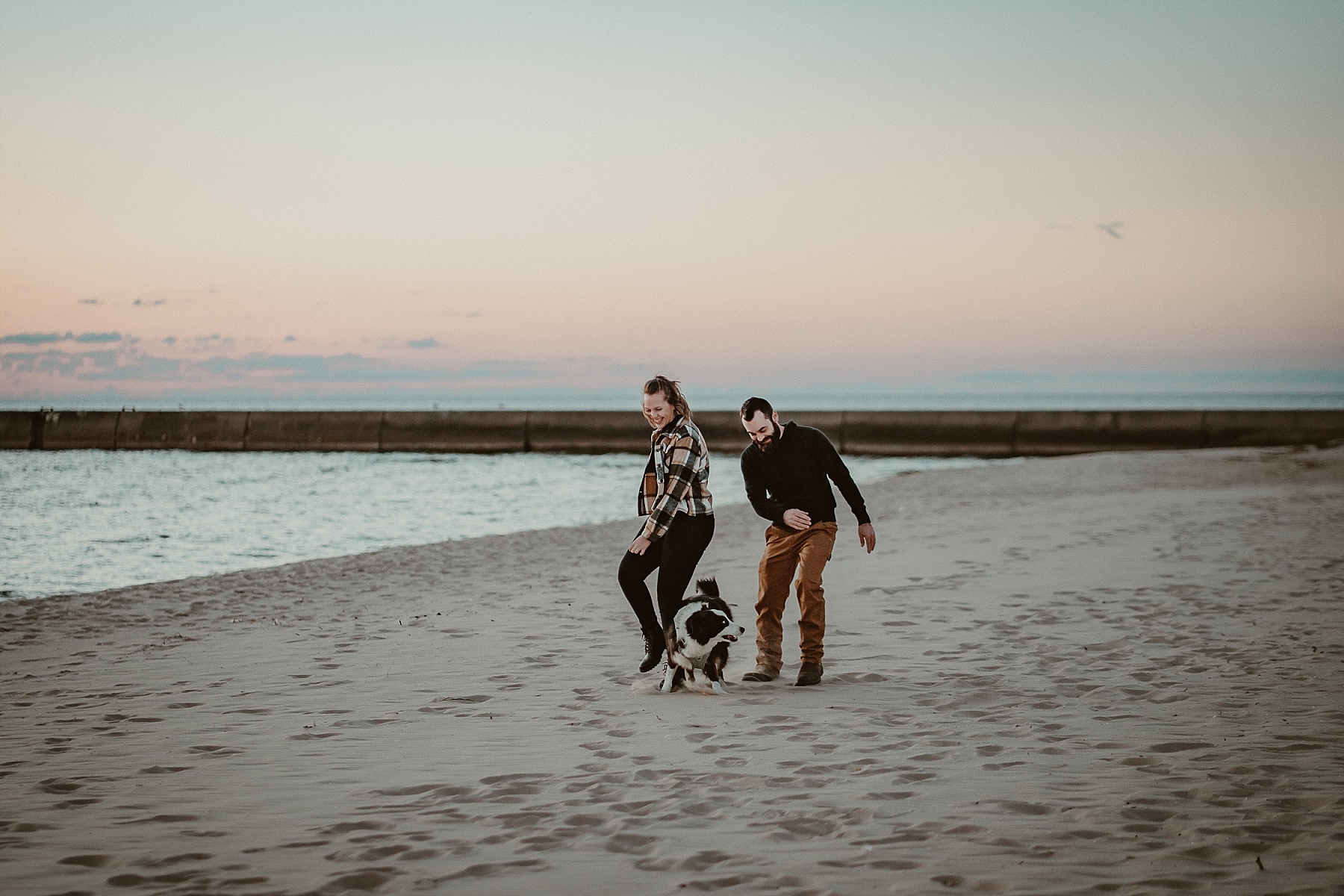 Border Collie and couple running along the shoreline during a Frankfort, Michigan engagement session.
