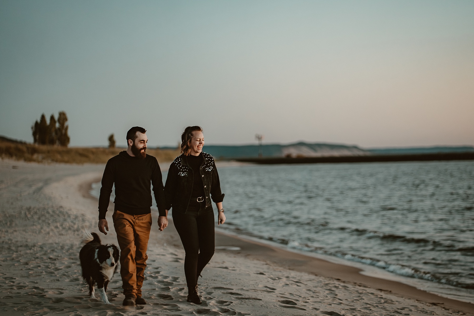 Couple holding hands walking with dog during a Lake Michigan sunset with pastel skies and waves rolling in.
