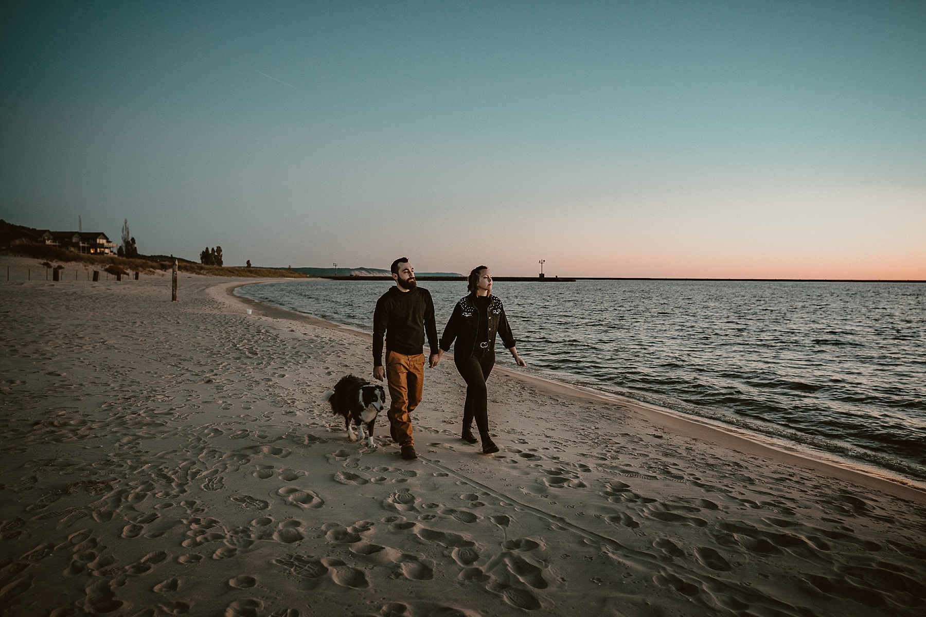 Couple holding hands walking with dog during a Lake Michigan sunset with pastel skies and waves rolling in.