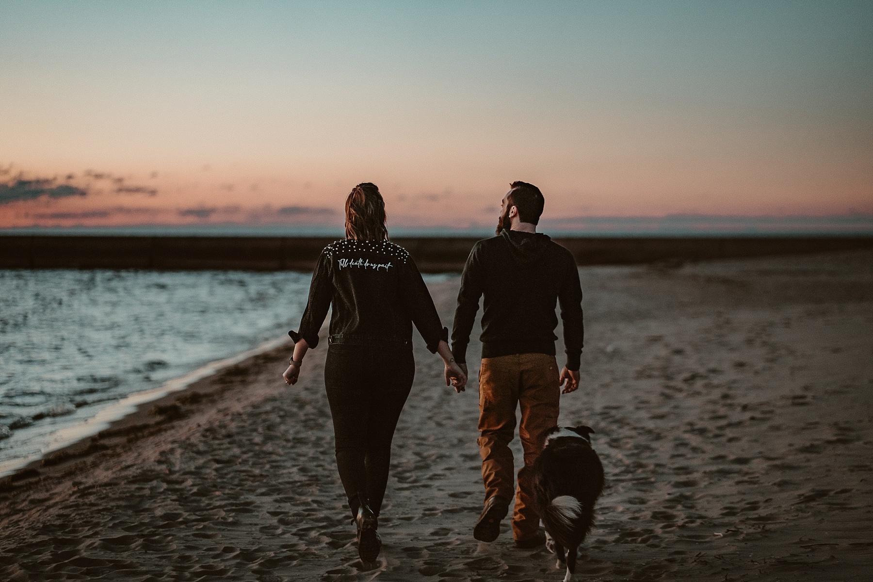 Couple with Border Collie holding hands walking during a Lake Michigan sunset with pastel skies.