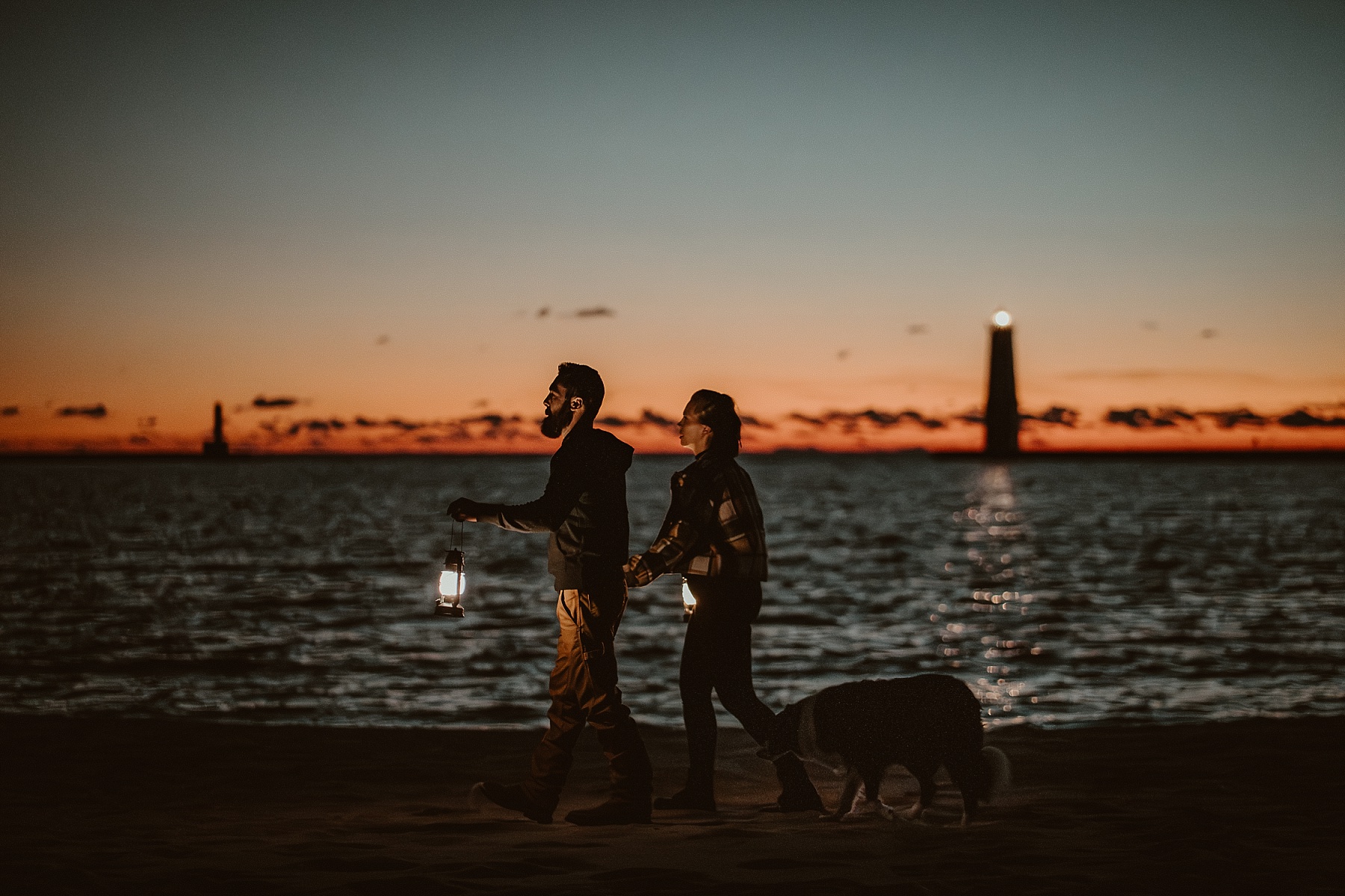 Engaged couple walking on the beach at sunset with lanterns and the Frankfort Light visible on the breakwater behind them.
