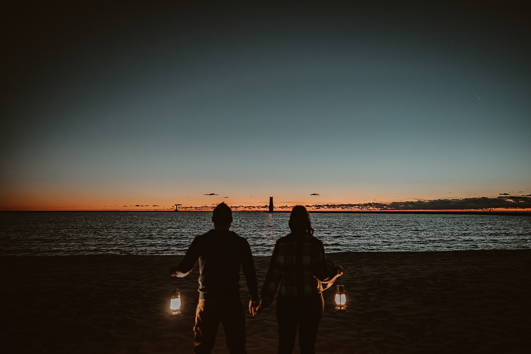 Engaged couple standing on the beach at sunset holding lit lanterns with Frankfort Light visible on the breakwater behind them.