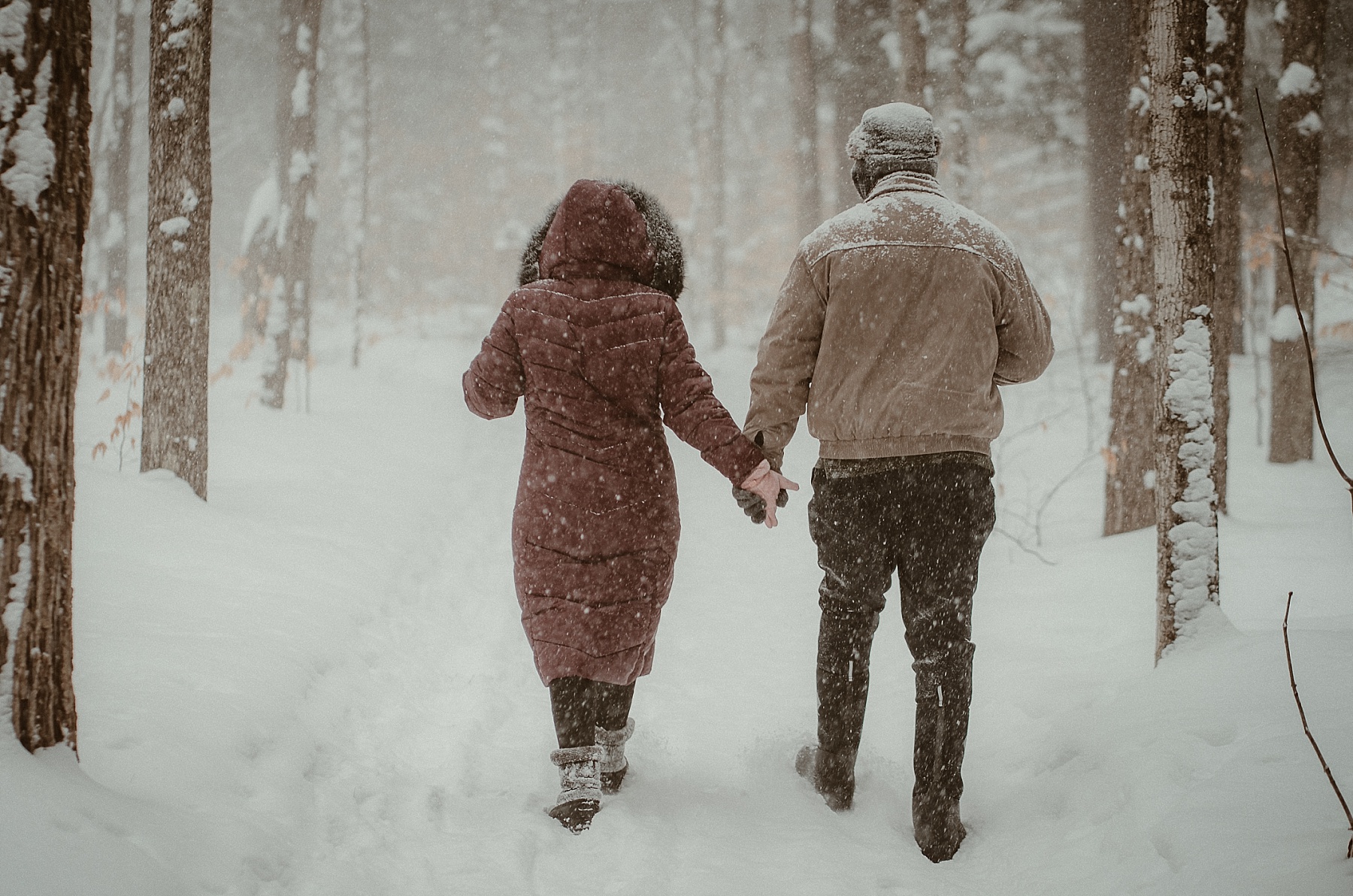 Couple walking through heavy snowfall during winter engagement session at Hartwick Pines State Park in Grayling, Michigan.