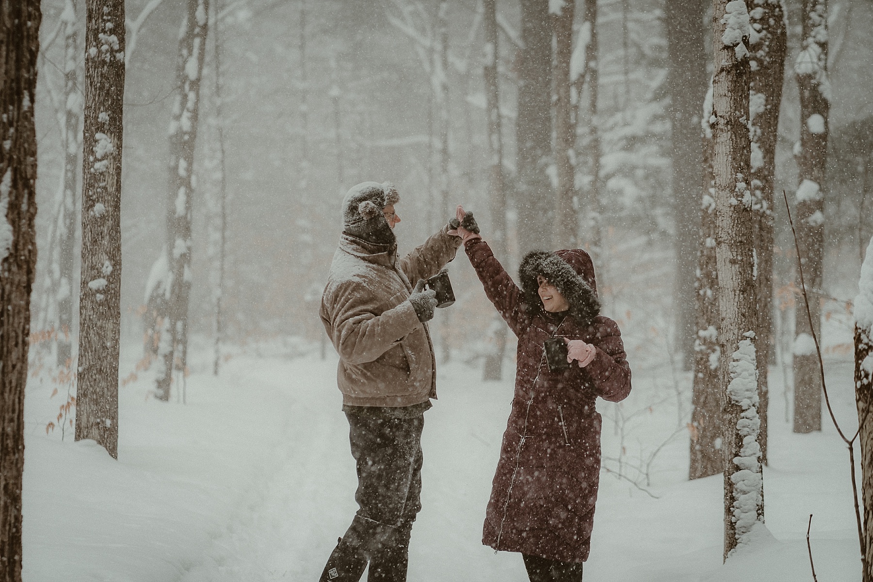 Couple laughing & dancing together in deep snow during cold-weather engagement session in Michigan.