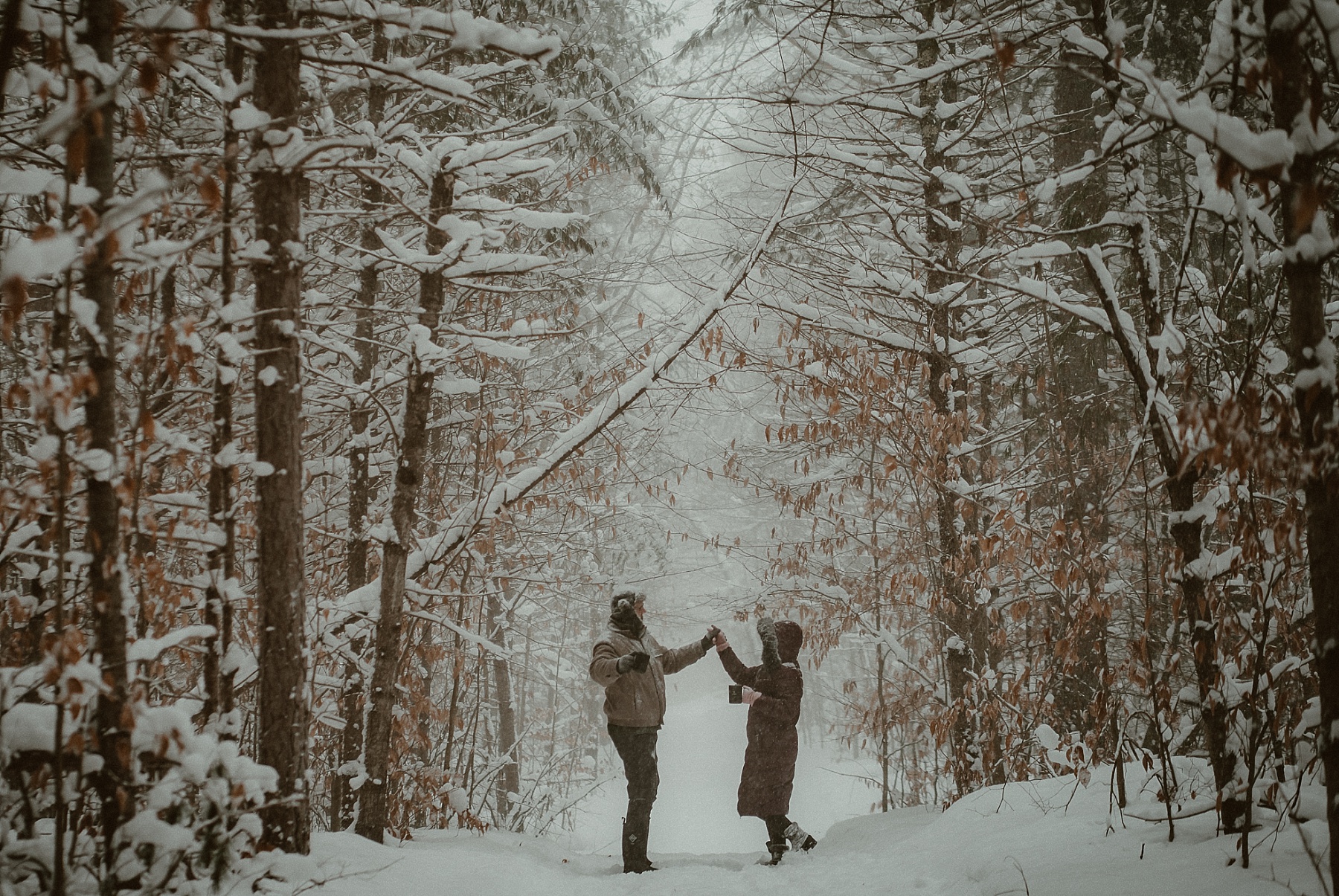 Couple laughing & dancing together in deep snow during cold-weather engagement session in Michigan.