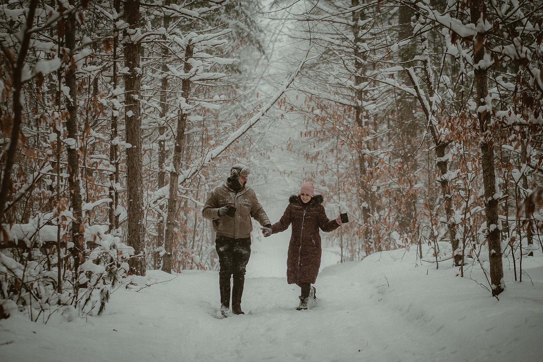 Couple walking & laughing through heavy snowfall during winter engagement session at Hartwick Pines State Park in Northern Michigan.