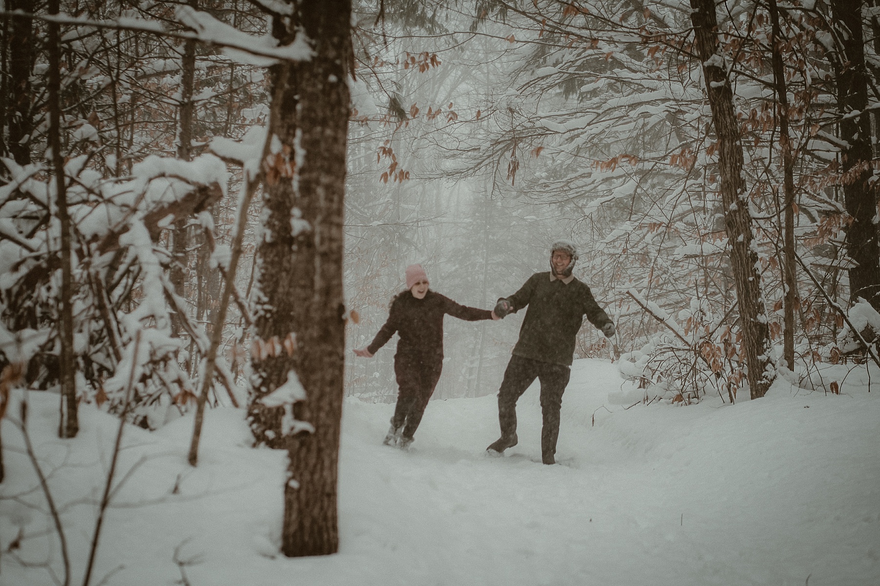 Couple running through heavy snowfall during winter engagement session at Hartwick Pines State Park in Northern Michigan.