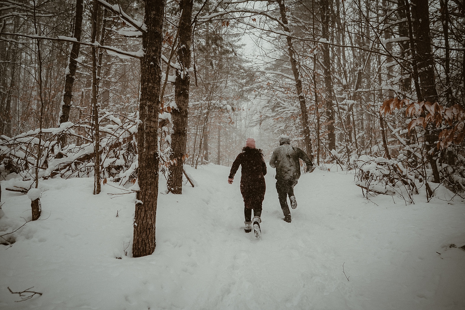 Couple running through heavy snowfall during winter engagement session at Hartwick Pines State Park in Northern Michigan.