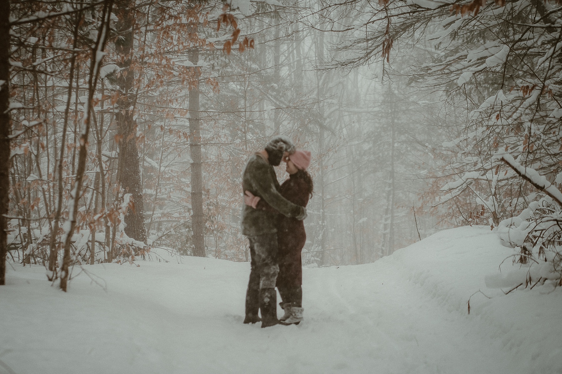Florida couple embracing in snowy pine forest during Northern Michigan winter engagement session.