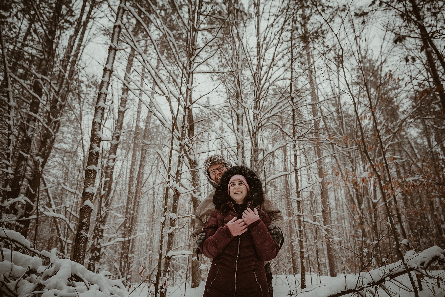 Florida couple embracing in snowy pine forest during Northern Michigan winter engagement session.