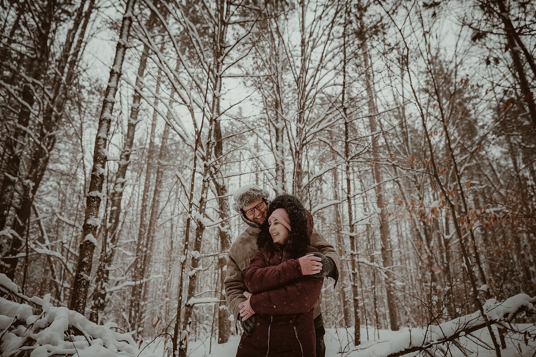Florida couple embracing in snowy pine forest during Northern Michigan winter engagement session.