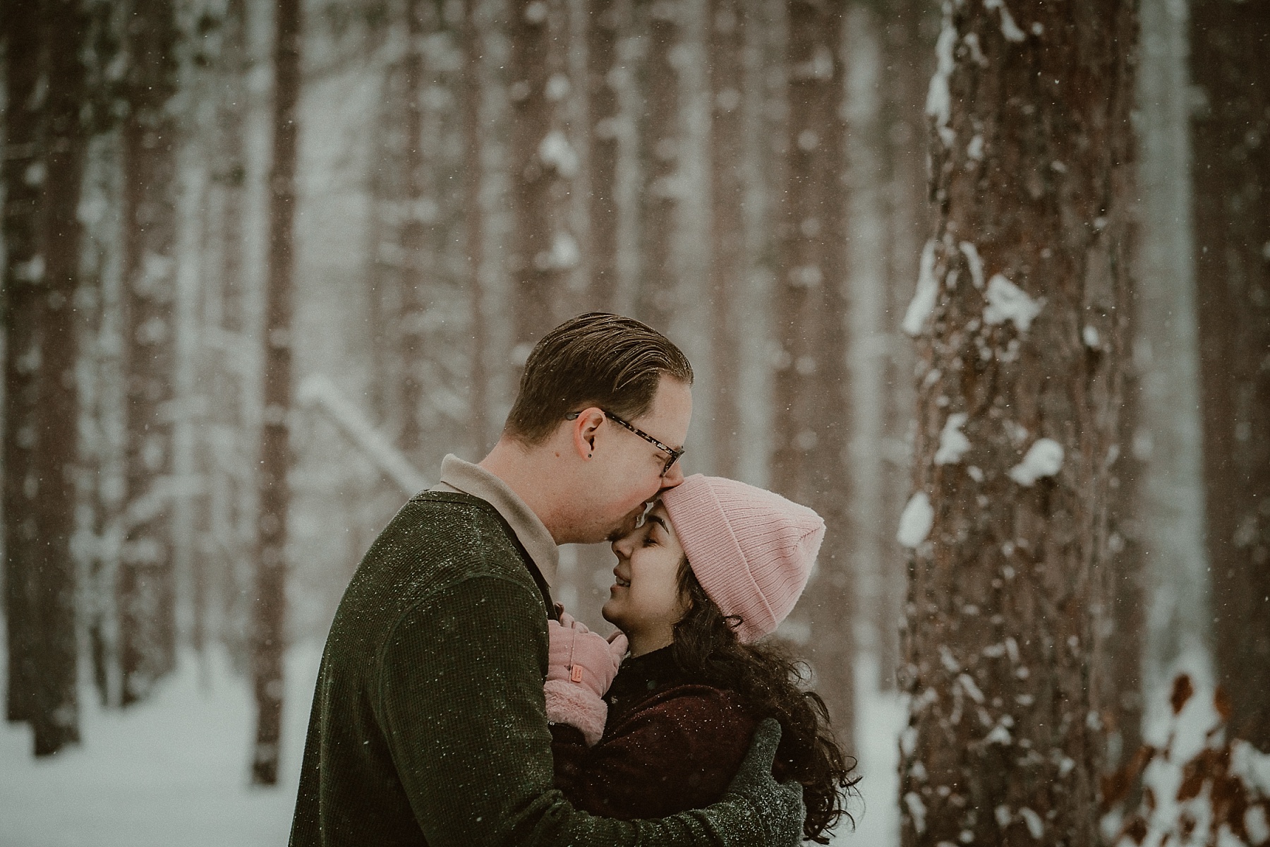 Florida couple embracing in snowy pine forest during Northern Michigan winter engagement session.