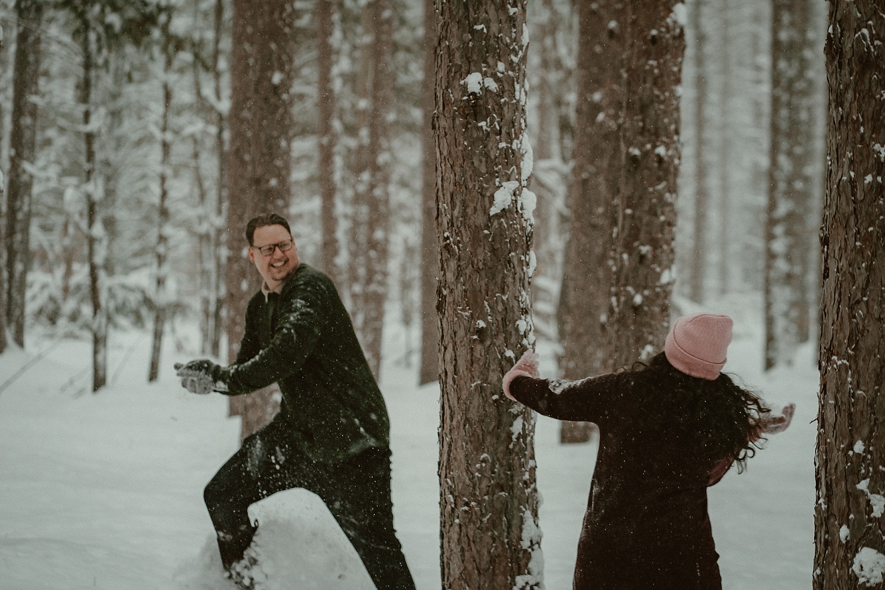 Couple having a snowball fight in heavy snowfall during winter engagement session at Hartwick Pines State Park in Grayling Michigan.