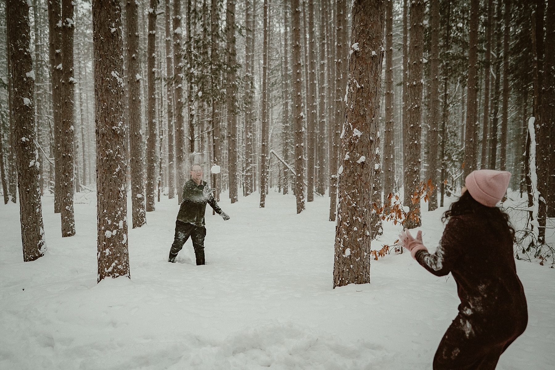 Couple having a snowball fight in heavy snowfall during winter engagement session at Hartwick Pines State Park in Grayling Michigan.