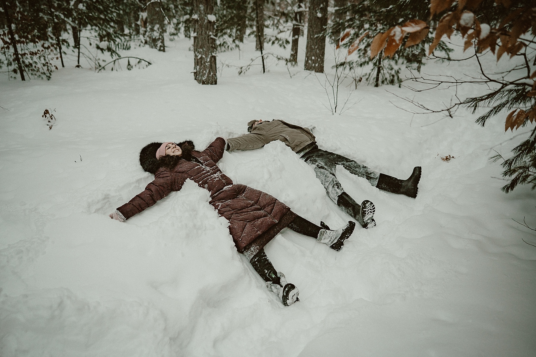 Couple making snow angels in heavy snowfall during winter engagement session at Hartwick Pines State Park in Grayling Michigan.