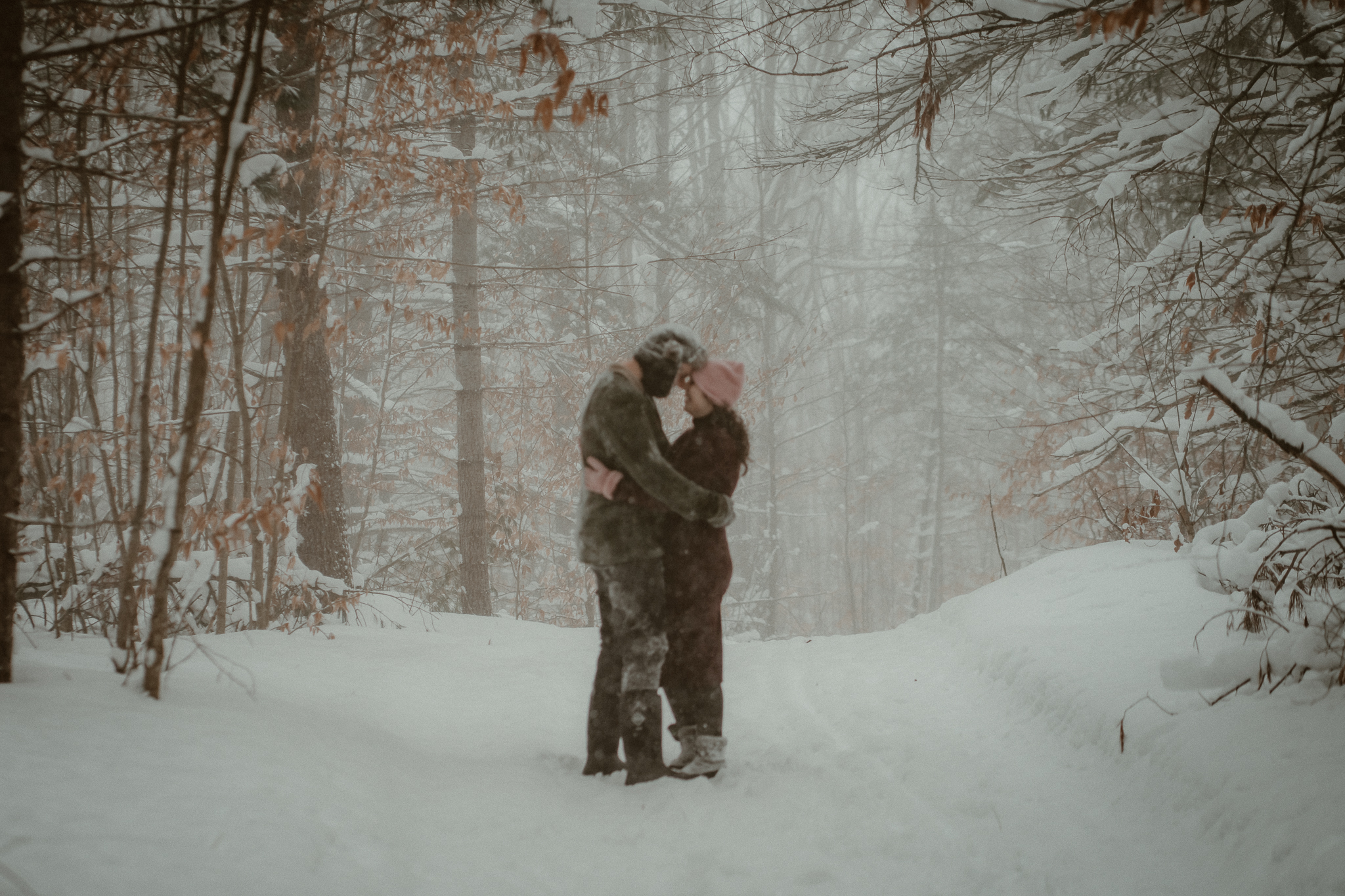 Florida couple embracing in snowy pine forest during Northern Michigan winter engagement session.